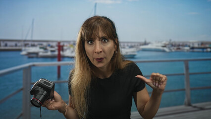 Young caucasian woman holding compact camera and thumb to chest by a pier railing on a street; self assertion defiance.