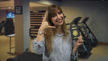 Woman smiling and holding a mexico passport while making a call me hand gesture inside a gym...