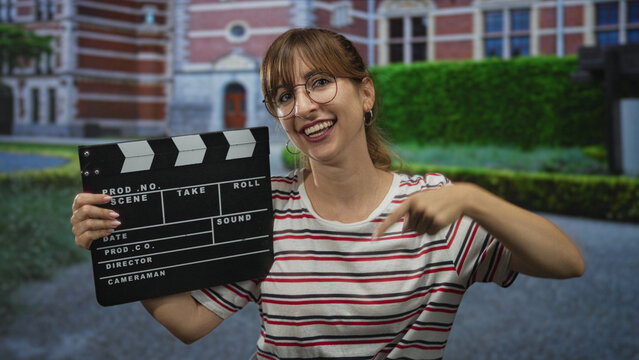 Woman holding clapperboard and pointing finger to clapperboard in front of historic building; filmmaking joy.
