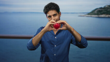 Man forms heart with hands holding red heart prop on building balcony overlooking sea; love affection warmth.