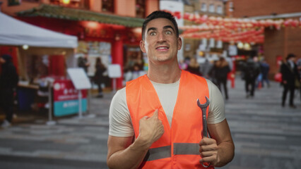 Man technician points finger to chest holding a wrench and wearing orange safety vest on busy street market with lanterns and stalls; pride responsibility.