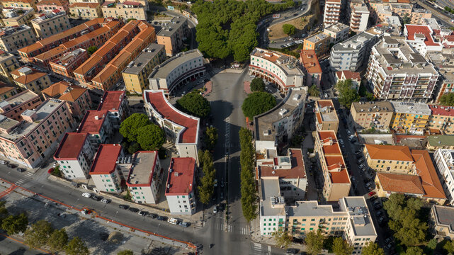 High-angle aerial view of a symmetrical urban plaza. Curving residential buildings with red roofs frame a central street, showcasing unique rationalist architecture and city planning.