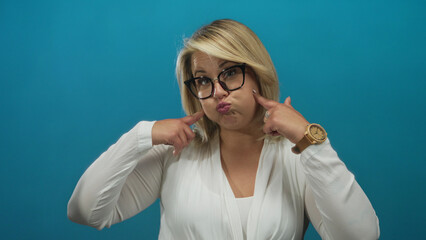 Woman with blonde hair and glasses wearing white blouse pointing fingers to cheeks in studio; playful amusement.