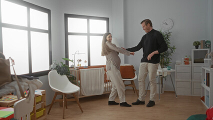 Pregnant couple dancing joyfully in bright nursery with crib, enjoying playful moment in cozy home, woman's hand on belly showing anticipation and excitement.