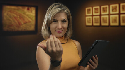 Young blonde woman taps tablet using interactive digital guide while standing before framed paintings in gallery space; focus.