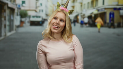 Fototapeta premium Woman with blonde hair wearing unicorn diadem smiles playfully outdoors on a busy city street with pastel buildings, creating a whimsical and joyful urban atmosphere.