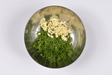 A top down view of freshly chopped green dill herb and minced garlic cloves combined in a stainless steel bowl on a white background