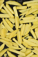 A top down view of many parboiled yellow potato slices spread across a black metal baking tray ready to be roasted in the oven
