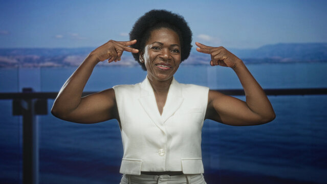 Woman points fingers to temples showing bare biceps on building balcony overlooking sea; confidence thinking.
