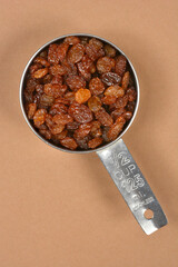 A top down view of a stainless steel measuring cup filled with sweet brown raisins placed on a plain brown background
