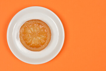 A top down view of a single round glazed candied orange slice served on a small white plate over a bright orange background with copy space