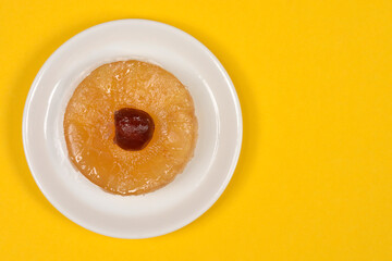 A top down view of a round glazed candied pineapple slice topped with a dark cherry served on a small white plate over a bright yellow background