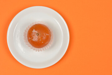 A top down view of a whole glazed candied clementine in a paper liner served on a small white plate over a bright orange background