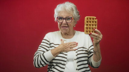 Elderly woman holds waffle in raised right hand with left hand on chest and smiles in studio;...