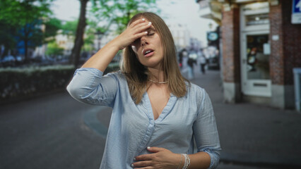 Fototapeta premium Woman clutching chest with one hand and pressing her stomach with the other while standing on a busy outdoor city street; distress.