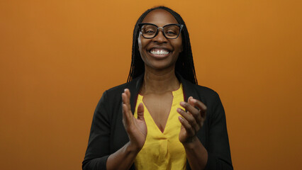 Woman clapping against orange background wearing yellow blouse and glasses, displaying joyful...