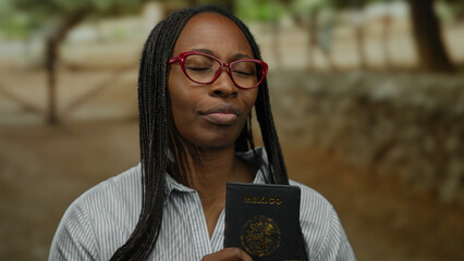 Woman outdoors holding a mexican passport while standing in a park setting with eyes closed, wearing red glasses and striped shirt signifying travel or immigration.