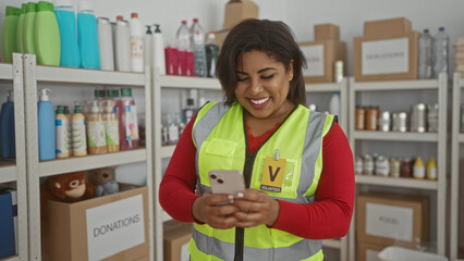 Woman volunteer wearing a reflective vest uses her phone in a charity donation center room with...