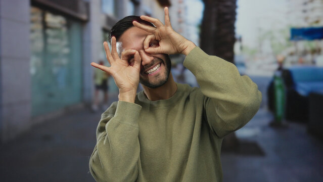 Hispanic man making glasses gesture with hands, smiling in an urban street setting, capturing a playful and humorous moment outdoors in a city environment.