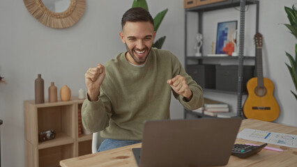 Hispanic man dancing joyfully in a cozy living room, celebrating while interacting with a laptop, surrounded by a modern home interior with a guitar and shelves.