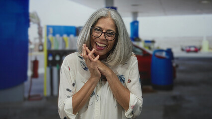 Middle age grey haired woman clapping hands on street near petrol station, smiling with glasses and open palms; joy.