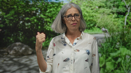 Woman with gray hair and glasses raising a clenched fist on a forest path while wearing a white...