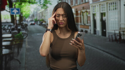 Fototapeta premium Young hispanic woman holding smartphone, finger to temple for headache while wearing brown tank top on street; frustration mobile stress.
