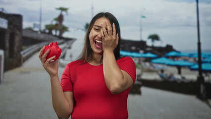Woman laughing and holding red piggy bank while covering one eye with hand on street by the beach...