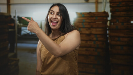 Woman points finger in a building amid stacked wooden crates and smiles with exposed arm; happiness playful.