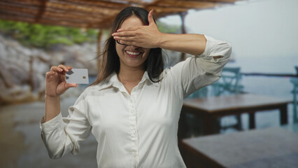 Woman holding creditcard and showing chip at seaside terrace with white shirt and glasses, raised arm gesture and visible hand; confidence trust finance.