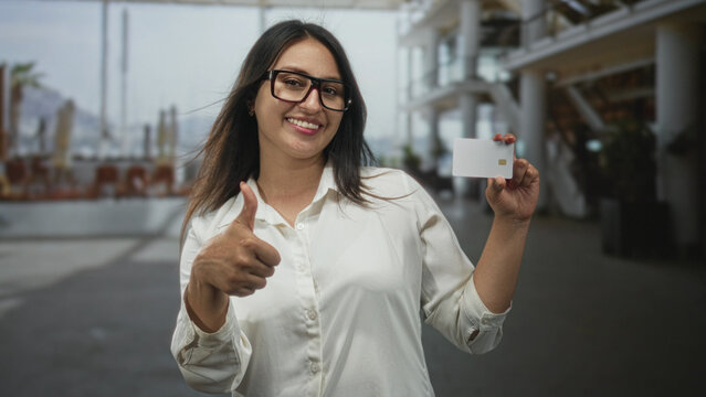 Woman gives thumbs up while holding a white creditcard with visible chip in building corridor, smiling at camera; confidence trust approval.