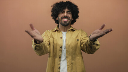 Man with hands to mouth blowing a kiss in studio with peach backdrop and mustard jacket, curly hair visible; playful affection.