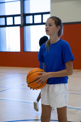 Teenage basketball teammates standing on gym court holding basketball next to tall windows