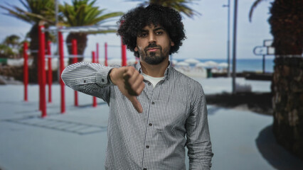 Man with curly hair showing thumbs down gesture at outdoor gym street with red fitness bars and palm trees; disapproval.