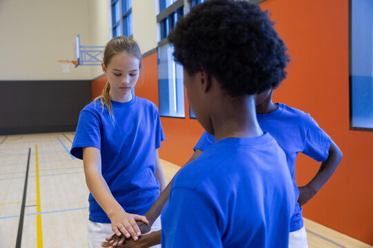 Diverse preteen teammates wearing shirts putting hands together in gym by basketball hoop