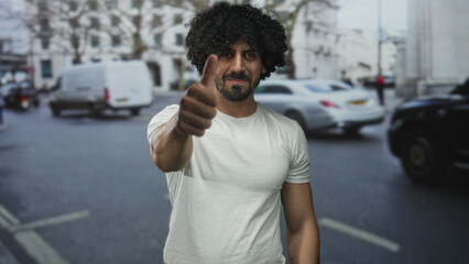 Man with curly hair showing thumbs up toward camera on a busy city street near parked cars and trees wearing white tshirt; confidence approval.