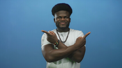Young african american man pointing fingers outward with crossed arms in blue studio, wearing white shirt and beaded bracelets; playful defiance.