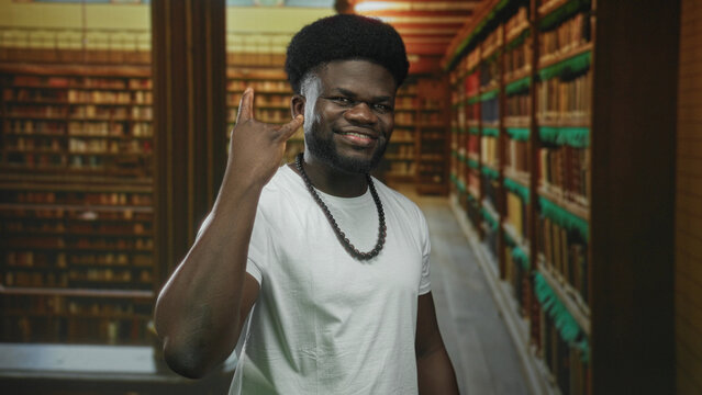 Man showing rock horns with right hand and smiling amid tall bookshelves in a library building aisle; confidence study focus.
