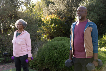 Senior couple in jackets exercising in garden lifting purple and black dumbbells