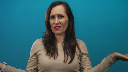 Obraz premium Woman with confused expression against blue background wearing beige sweater with long brown hair raising hands in questioning gesture capturing unsure emotion.