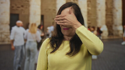 Fototapeta premium Woman in yellow sweater covering face on historic street with blurred people and ancient stone wall in background depicting outdoor urban scene in old town.