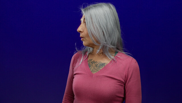 Senior grey-haired woman in a pink shirt looking surprised against isolated blue background, capturing candid emotional expression.