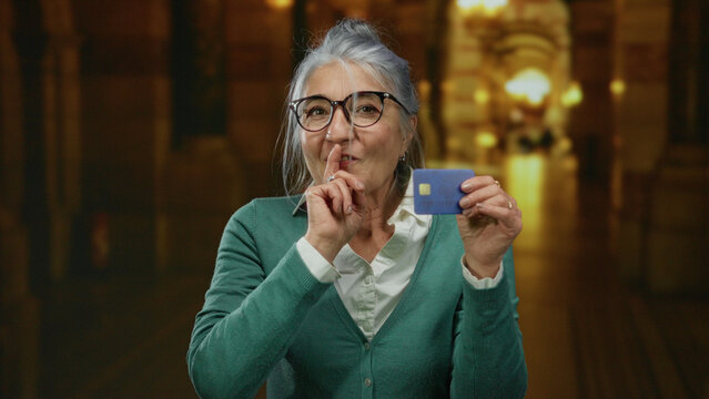 Senior woman with grey hair holding credit card in church while gesturing silence, wearing glasses and green sweater, indoor scene providing contrast and mystique.