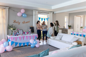 Diverse female friends standing and chatting in living room under baby shower banner, balloons
