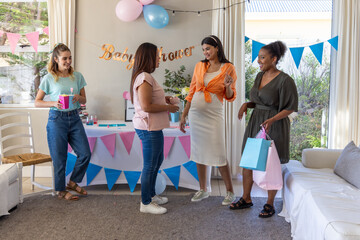 Diverse female friends celebrating baby shower in living room with gifts and balloons