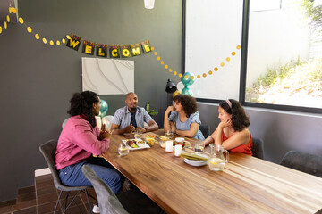 Diverse friends sitting around wooden dining table in gray-walled room, banner, fruit, garlands