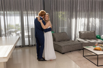Couple embracing in modern living room by sheer curtains with wine glasses and balloons