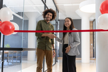 Diverse coworkers shaking hands across red ribbon in office lobby with balloons, glass partition © wavebreak3