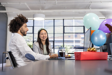 Diverse coworkers sitting at desk in open-plan office celebrating gift box, balloons by computer
