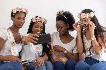 Diverse female friends wearing white tops taking selfie on living room sofa with smartphone, sashes